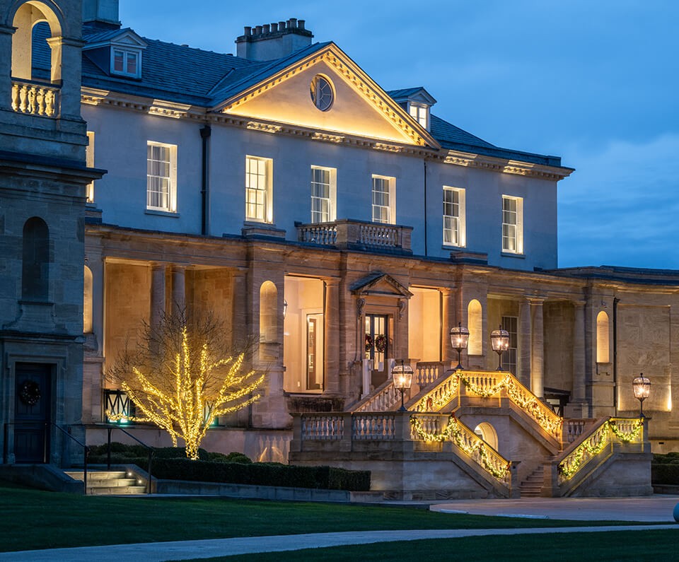 The Langley, an elegant mansion exterior at dusk with warm lights illuminating the facade and steps. A tree adorned with glowing fairy lights enhances the inviting ambiance.